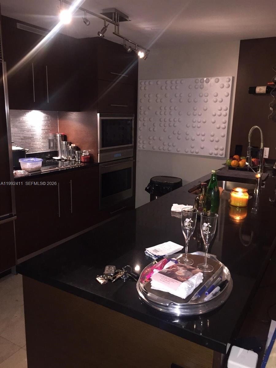 A modern kitchen features dark wood cabinetry, black countertops, stainless steel appliances, and track lighting — 465 Brickell Ave # 5605, Miami FL 33131, MIAMI, FL