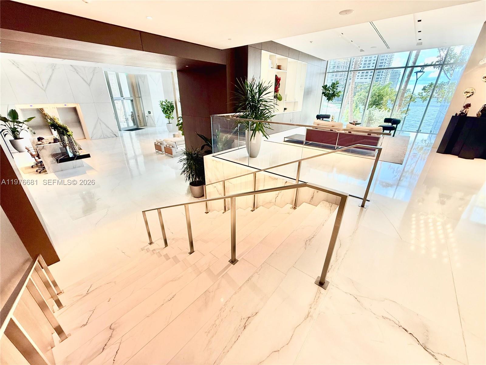A modern lobby features marble flooring, a glass-railed staircase, and floor-to-ceiling windows overlooking water — 300 Biscayne Boulevard Way # 3608W, Miami FL 33131, MIAMI, FL