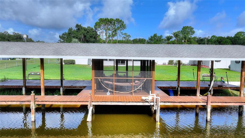 A screened-in wooden deck overlooking a lake features a dining set, chairs, and a hammock — 6197 AMARYLLIS DRIVE, INDIAN LAKE ESTATES, FL