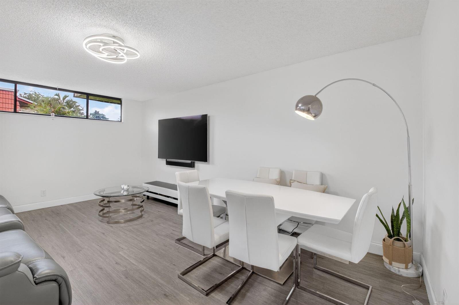 A modern dining and living area features white furniture, a wall-mounted television, and wood flooring — 8385 Trent Court # D, Boca Raton FL 33433, BOCA RATON, FL