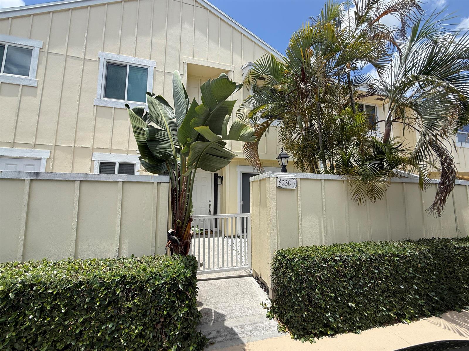 A beige two-story townhouse features a gated courtyard entrance, a large plant, and palm trees — 6238 Riverwalk Lane # 7, Jupiter FL 33458, JUPITER, FL