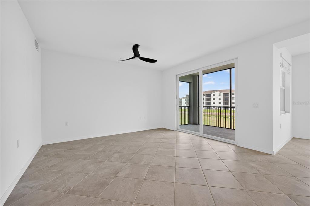 A bright, empty living room features large beige floor tiles, a ceiling fan, and sliding doors — 18461 COPPERHEAD COURT N UNIT 422, LEHIGH ACRES, FL