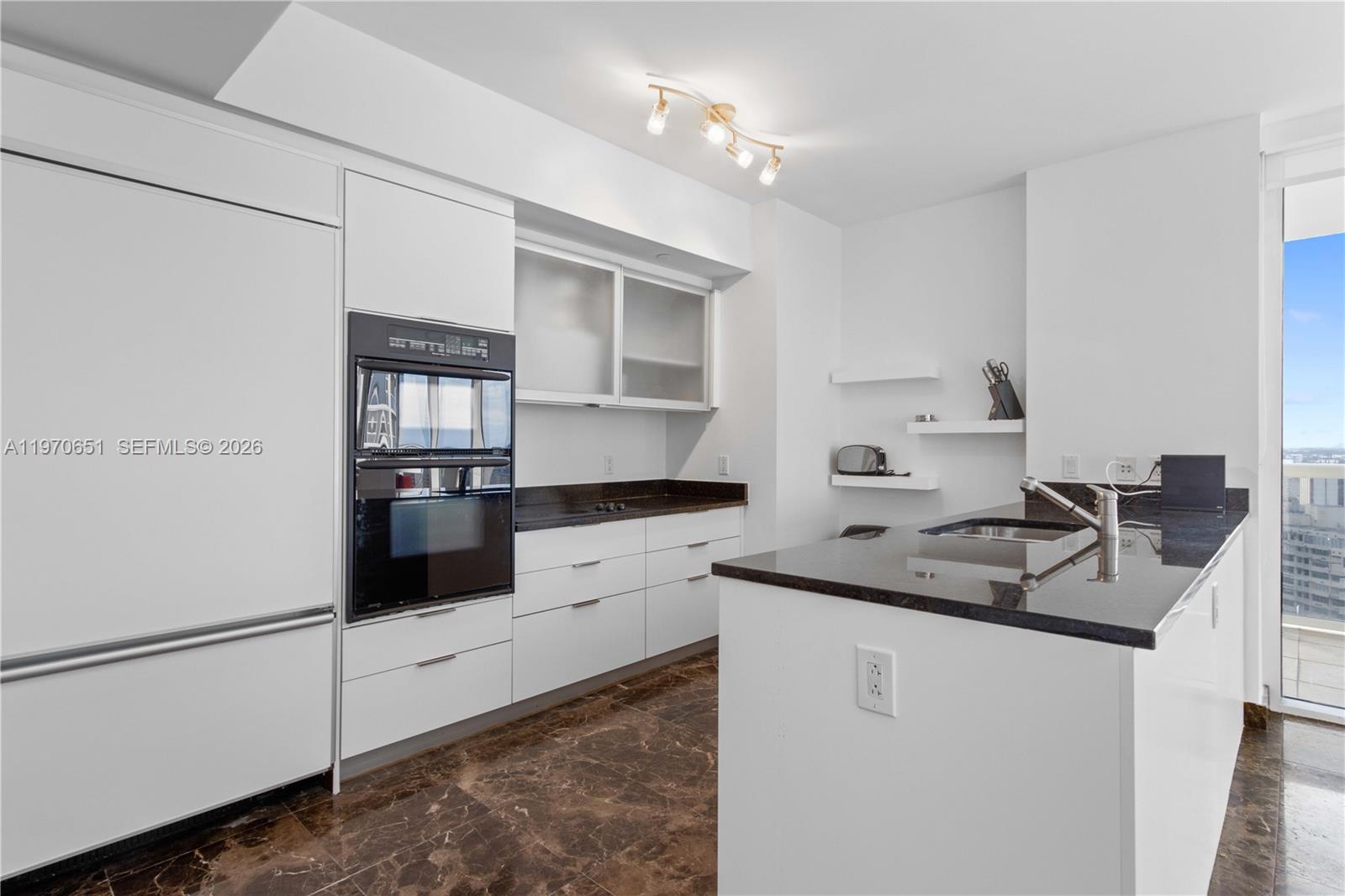 A modern kitchen features white cabinetry, dark granite countertops, and brown marble flooring — 1830 S Ocean Dr # 1908, Hallandale Beach FL 33009, HALLANDALE BEACH, FL