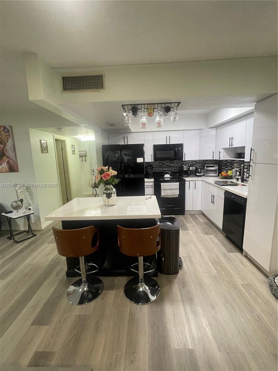 A kitchen features white cabinets, black appliances, a central island with bar stools, and wood-look flooring — 6945 W 2nd Way, Hialeah FL 33014, HIALEAH, FL