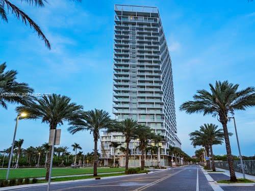 A tall modern residential high-rise building stands behind a palm-lined street at dusk — 2000 Metropica Way # 2610, Sunrise FL 33323, SUNRISE, FL