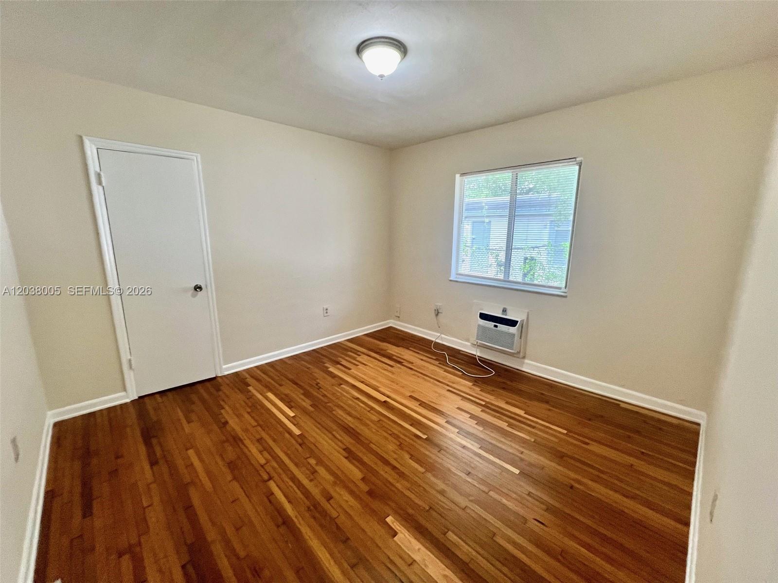 An empty bedroom features hardwood flooring, a white door, a window, and a wall-mounted air conditioner — 692 NE 85th St # 5, Miami FL 33138, MIAMI, FL