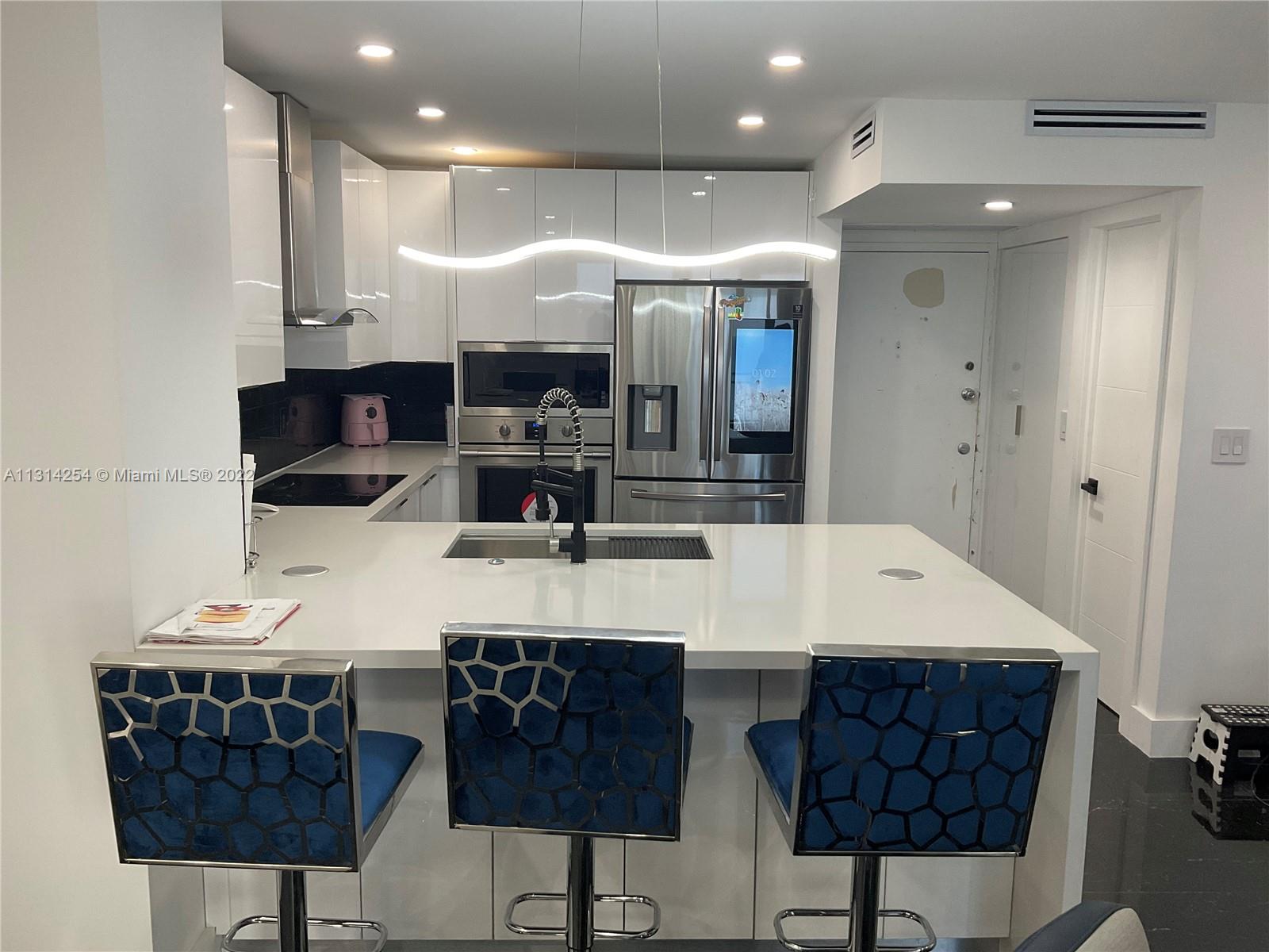 A modern kitchen features white cabinetry, stainless steel appliances, a quartz island, and blue stools — 3180 S Ocean Dr # 1503, Hallandale Beach FL 33009, HALLANDALE BEACH, FL