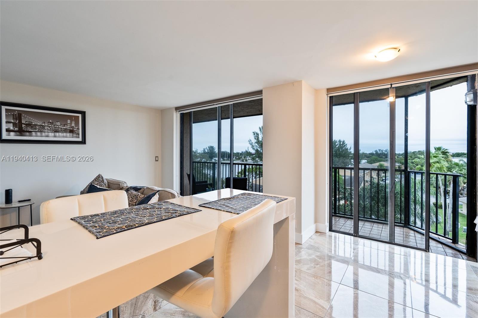 A dining area features a white table, modern chairs, marble floors, and sliding glass balcony doors — 3400 Blue Lake Dr # 505, Pompano Beach FL 33064, POMPANO BEACH, FL