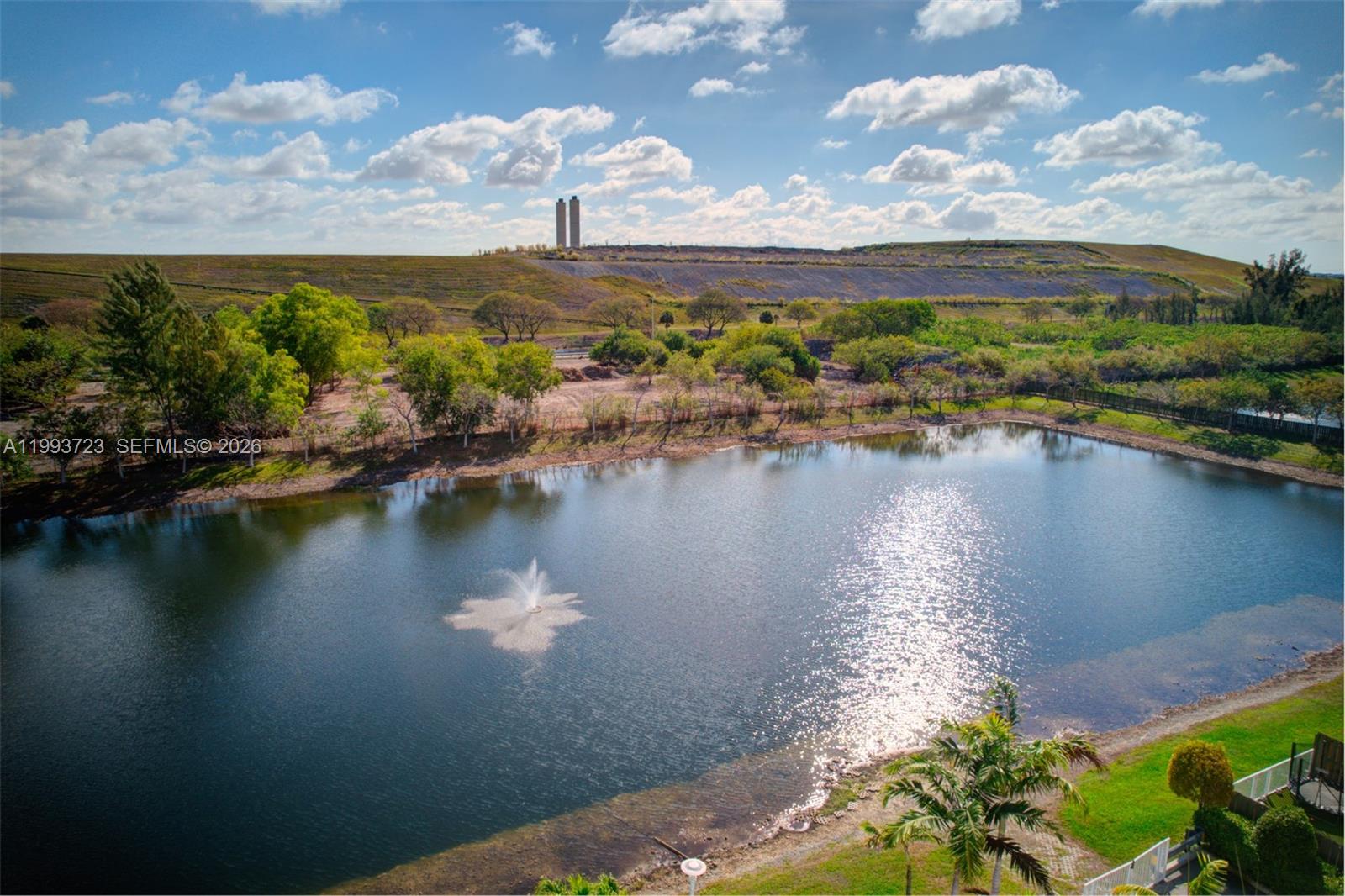 An aerial view shows a pond with a fountain, surrounding trees, and a distant hill — 7159 NW 102nd Place, Doral FL 33178, DORAL, FL
