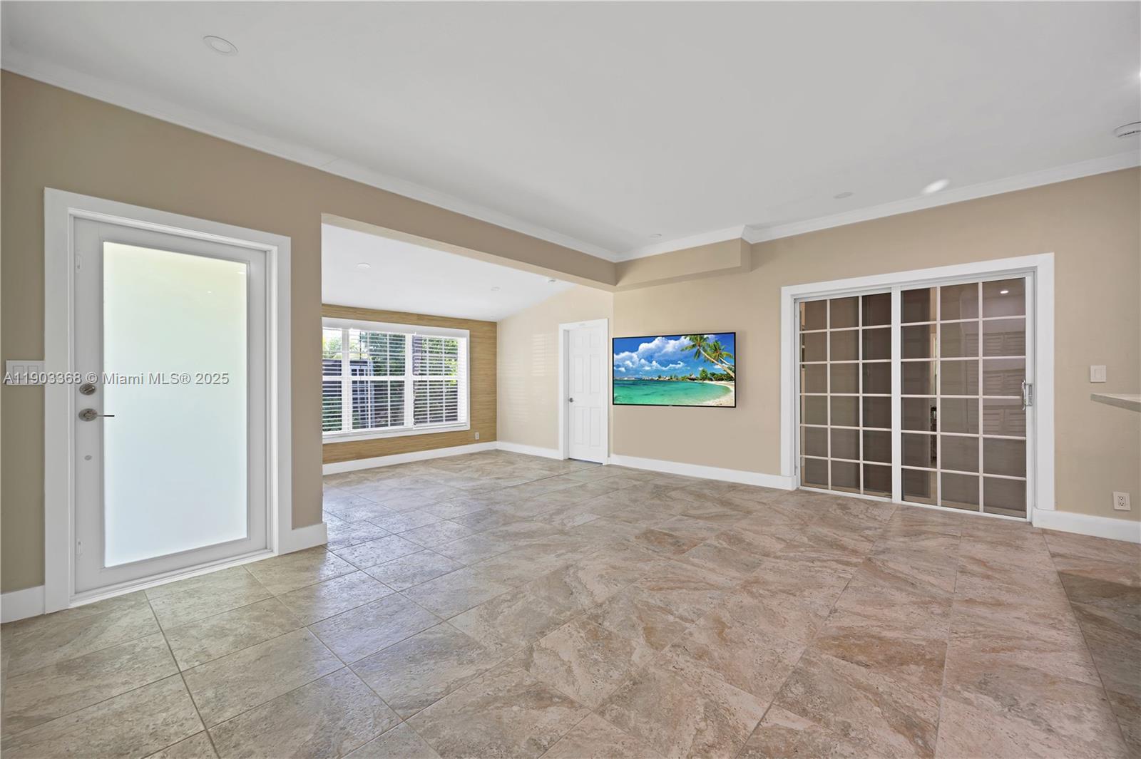 A spacious room features beige tile flooring, a wall-mounted television, and glass-paned sliding doors — 818 NE 3rd St, Hallandale Beach FL 33009, HALLANDALE BEACH, FL