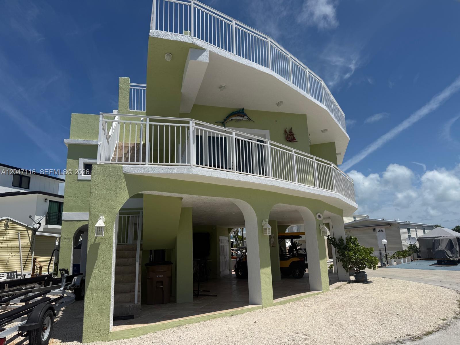 A light green, multi-story elevated house features white railings, arched supports, and a gravel driveway — 65821 Overseas Hwy 176, Long Key FL 33001, LONG KEY, FL