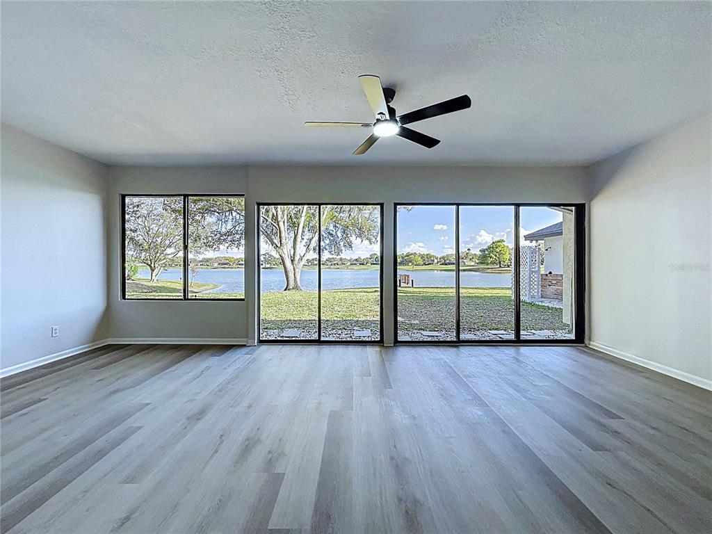An empty living room features light wood-look flooring, a ceiling fan, and large windows overlooking a lake — 2881 HAMMOCK DRIVE, PLANT CITY, FL