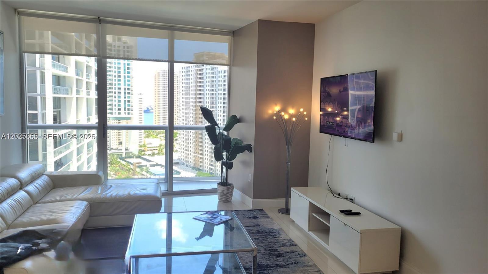 A living room features a white sofa, glass coffee table, wall-mounted television, and floor-to-ceiling windows — 485 Brickell Ave # 2311, Miami FL 33131, MIAMI, FL