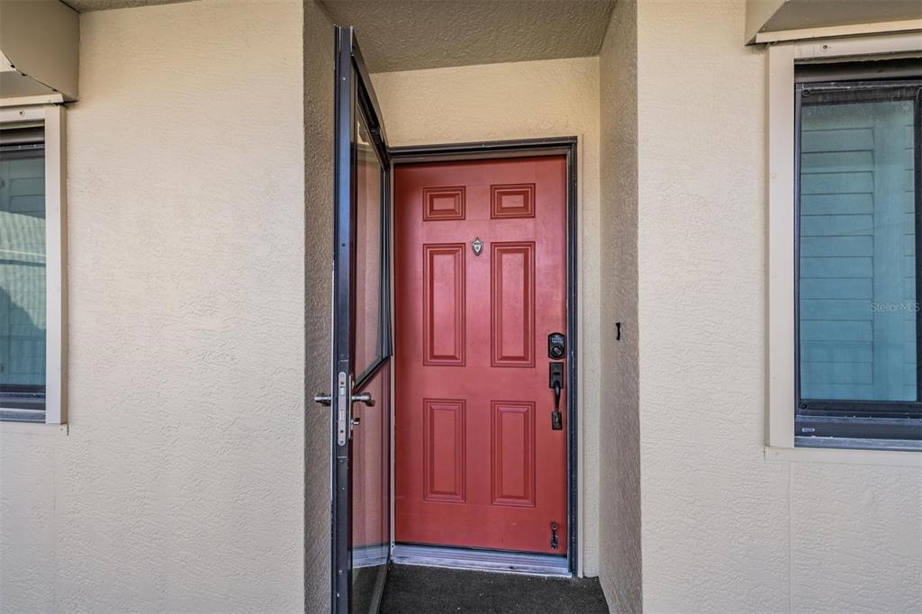 A red front door with a storm door is set between two windows on a stucco wall — 7435 BAYSHORE DRIVE UNIT 303, TREASURE ISLAND, FL