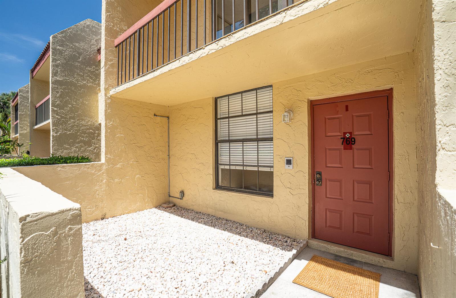 A tan stucco building exterior features a red front door, a gravel patio, and balcony — 769 SE 1st Way # 85, Deerfield Beach FL 33441, DEERFIELD BEACH, FL