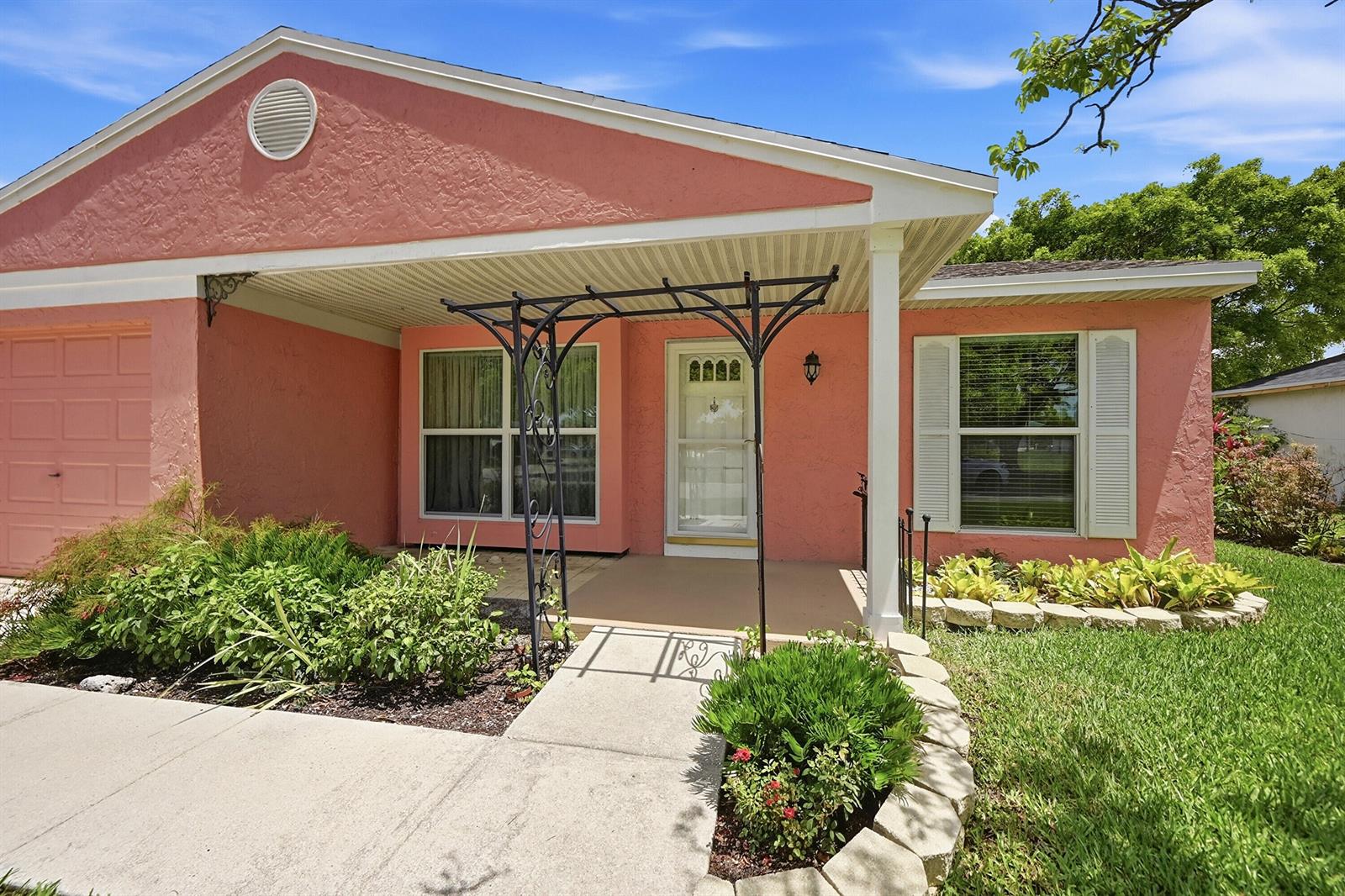 A pink stucco house features a covered entryway with a metal trellis and white shutters — 8131 Countrypark Drive, Boca Raton FL 33433, BOCA RATON, FL