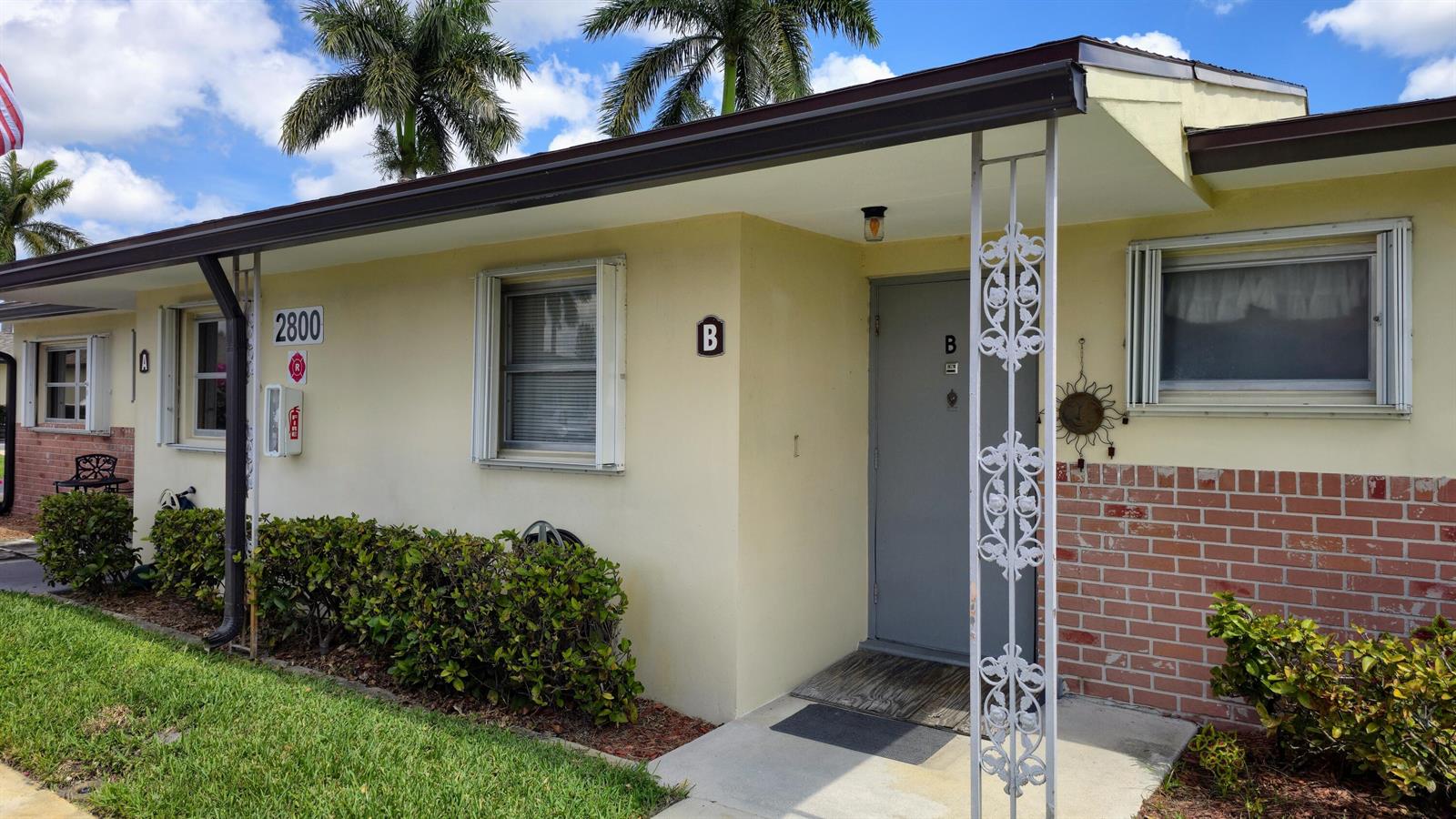The exterior of a single-story building features light yellow walls, a brick base, and unit B — 2800 Ashley Drive E # B, West Palm Beach FL 33415, WEST PALM BEACH, FL