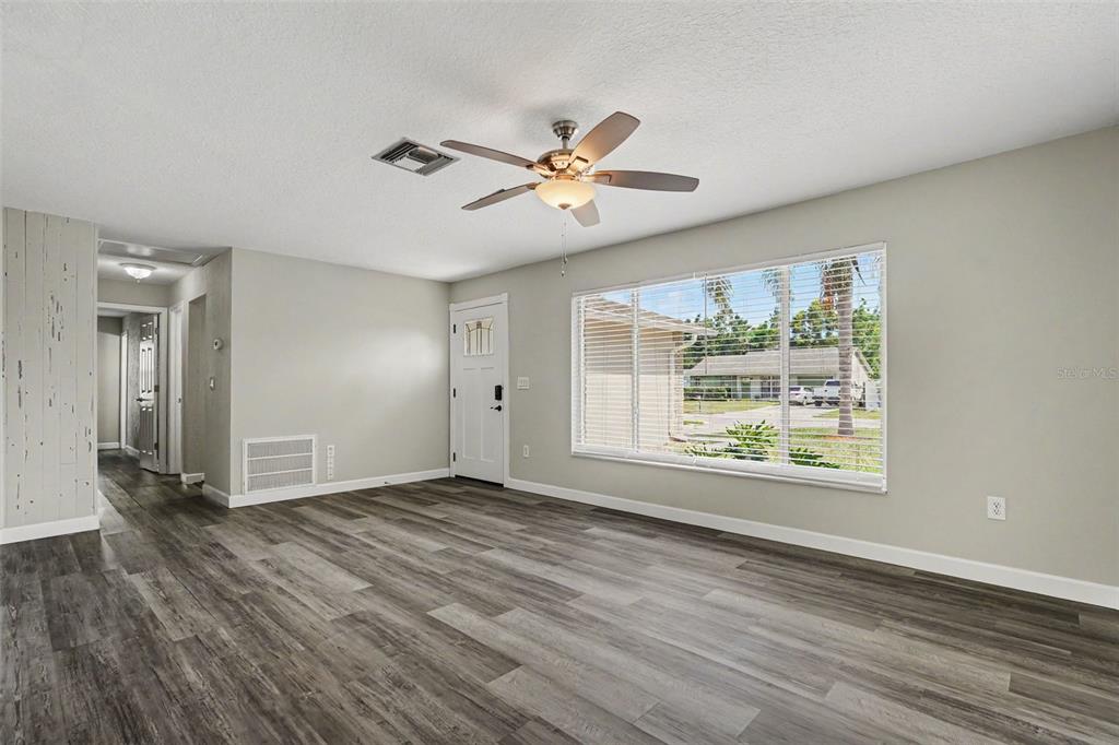 A living room features gray wood-look flooring, a ceiling fan, and a large front window — 6822 LASSEN AVENUE, NEW PORT RICHEY, FL