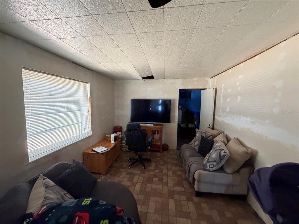 A living room features a wall-mounted television, a sofa, a gaming chair, and damaged ceiling tiles — 10904 ROSS ST, TAMPA, FL
