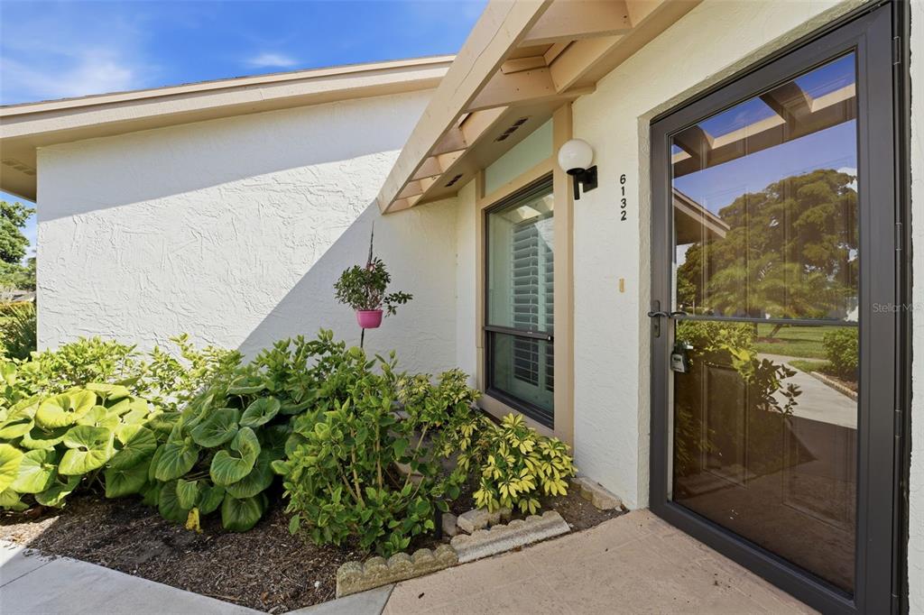 A white stucco house exterior features a dark-framed storm door and a landscaped garden bed — 6132 29TH AVENUE W UNIT 6132, BRADENTON, FL