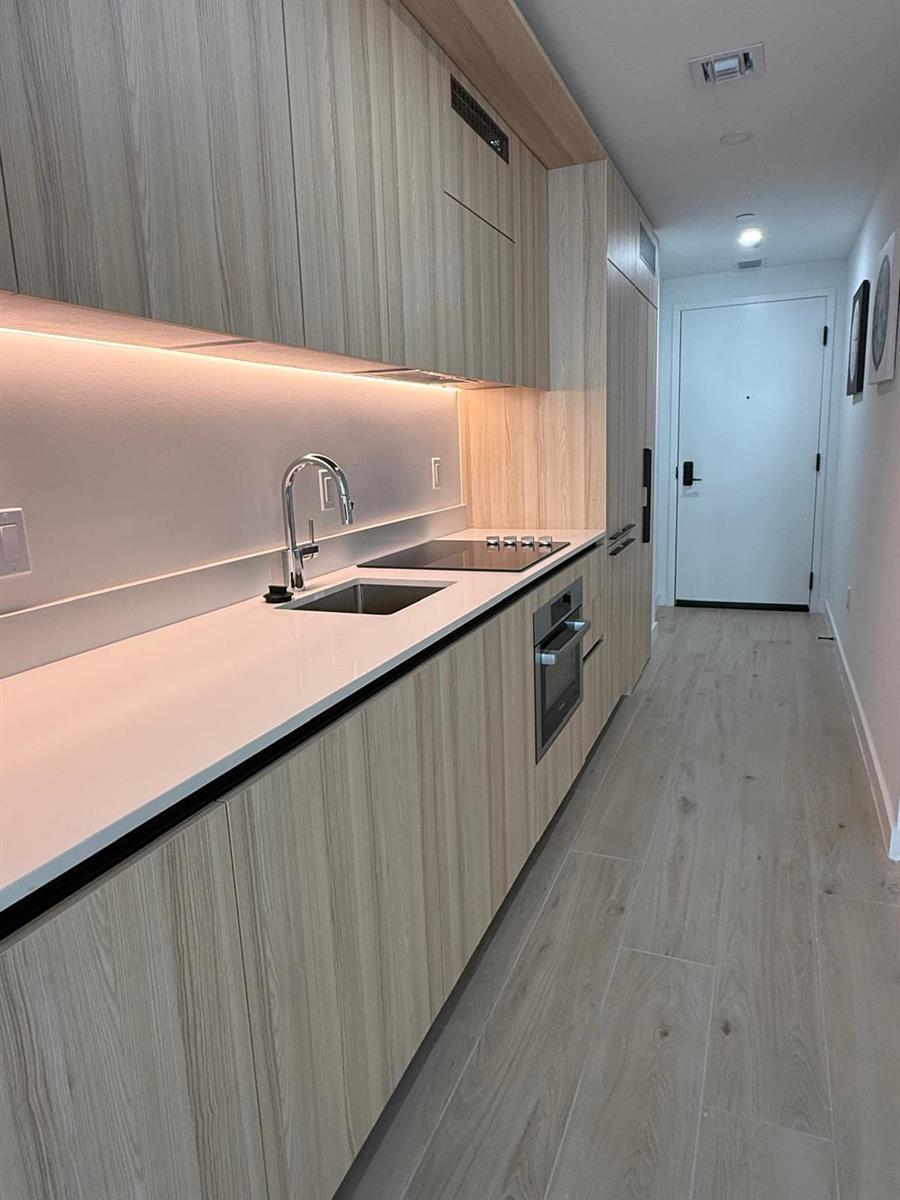 A modern galley kitchen features light wood cabinetry, white countertops, and integrated stainless steel appliances — 225 N Miami Avenue # 910, Miami FL 33128, MIAMI, FL