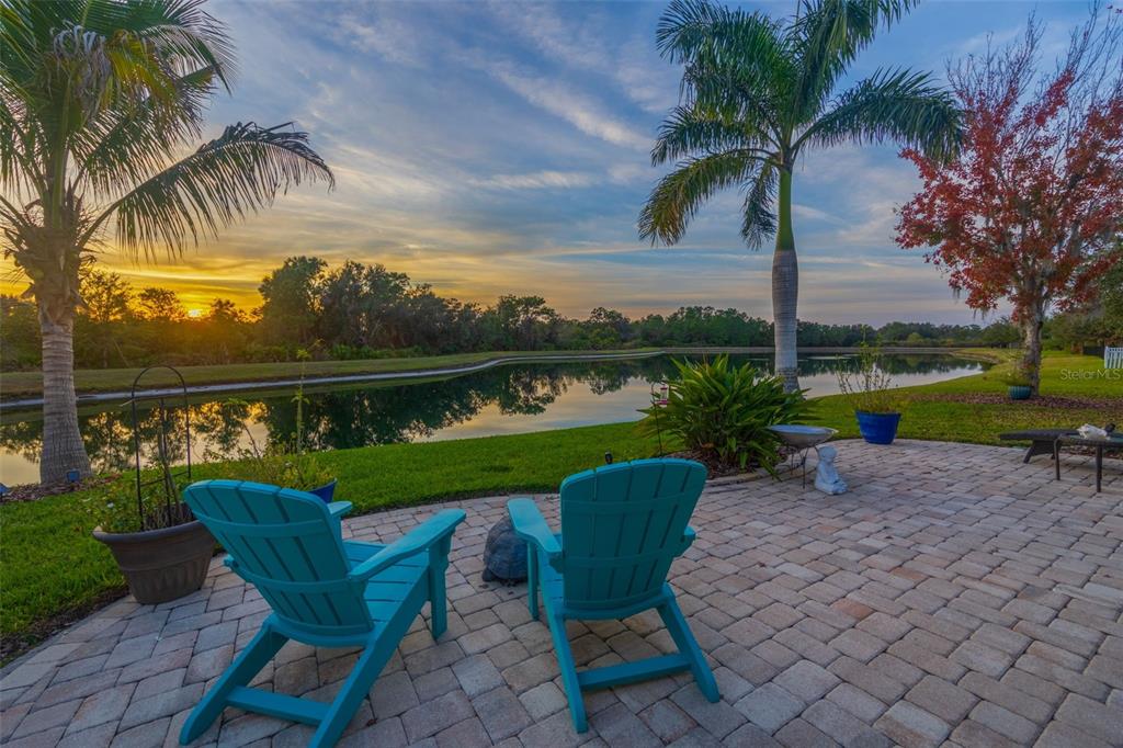 Two turquoise Adirondack chairs sit on a brick patio overlooking a pond at sunset — 5652 FISHER GLEN LOOP, WESLEY CHAPEL, FL
