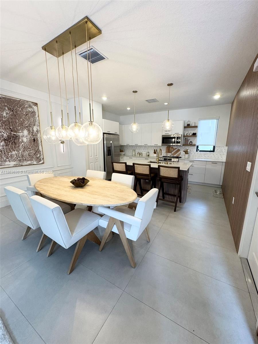 A modern dining area with an oval table and pendant lights opens into a white kitchen — 4867 NW 84th Ave, Doral FL 33166, DORAL, FL