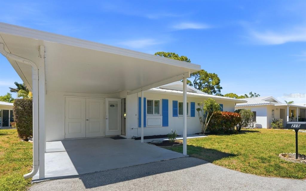 A white single-story house features a large carport, blue shutters, and a front lawn — 215 FAIR WINDS DRIVE UNIT 215, NOKOMIS, FL