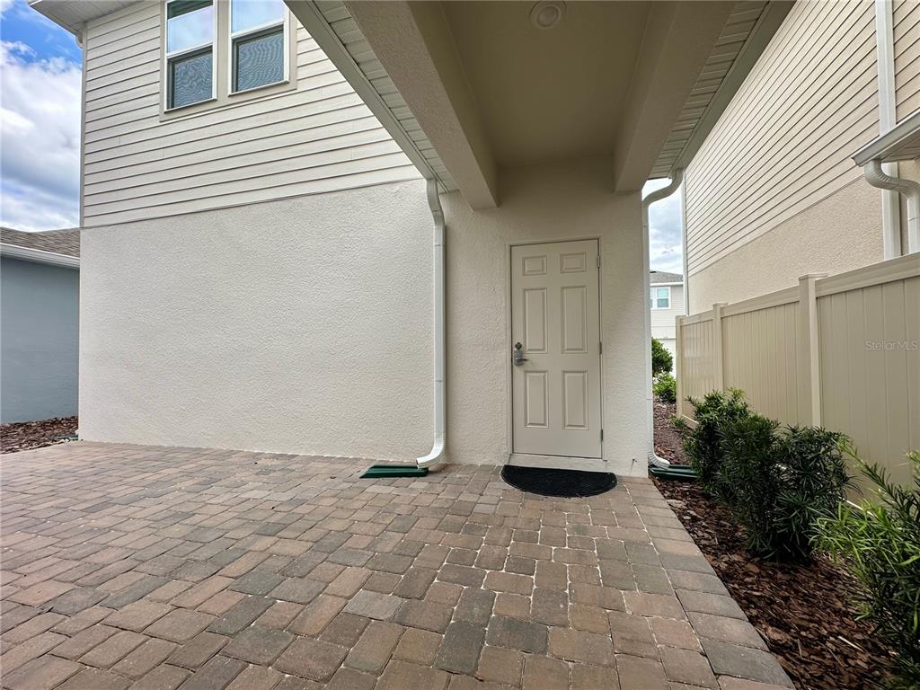 A paved patio sits beneath an overhang next to a beige door and vinyl fence — 12761 HARVEST RAIN AVENUE, WINTER GARDEN, FL