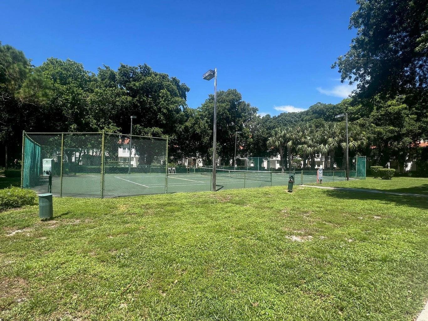A fenced outdoor tennis court sits on a grassy lawn under a clear blue sky — 1050 Crystal Way # D, Delray Beach FL 33444, DELRAY BEACH, FL
