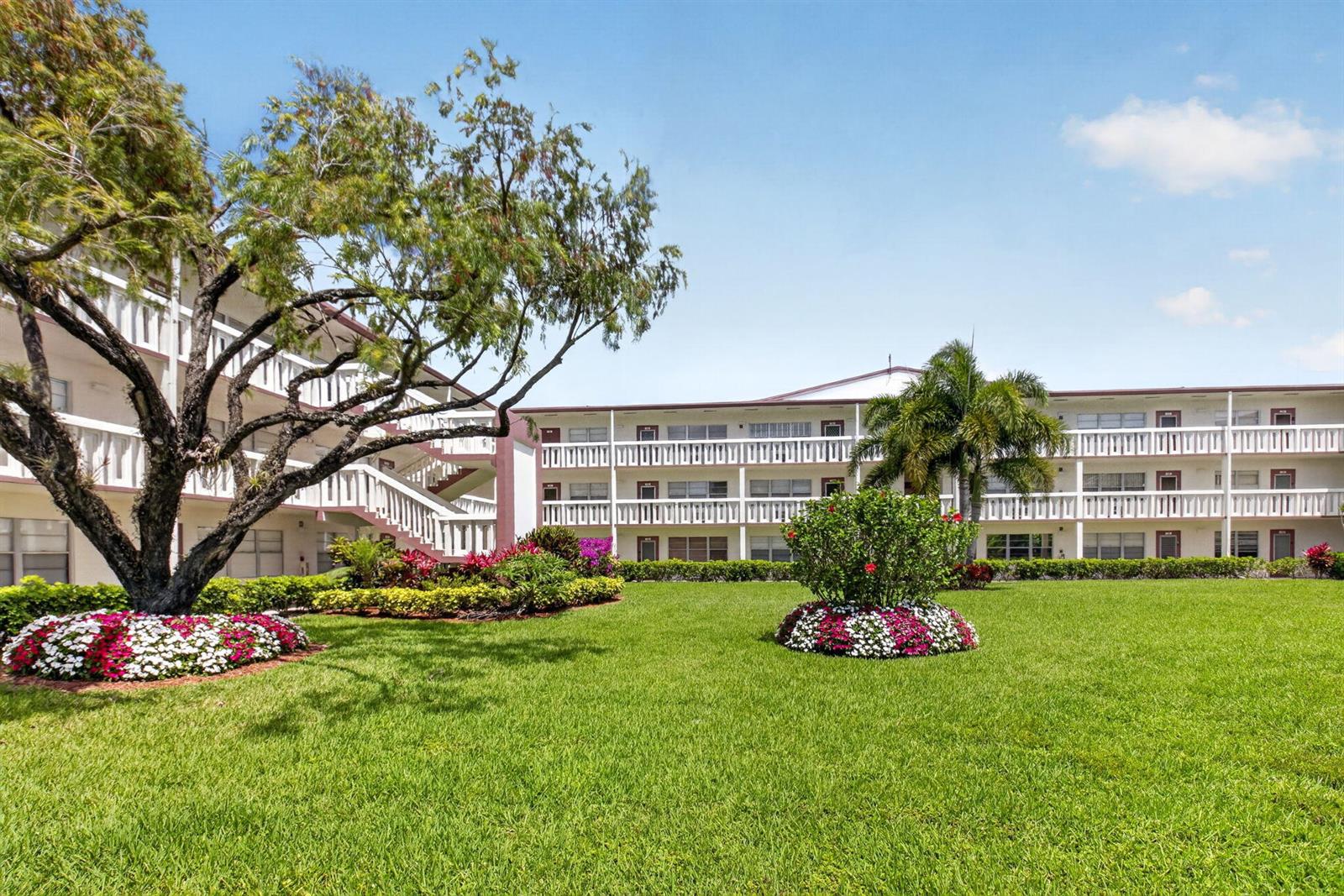 A multi-story apartment building with white balconies overlooks a grassy courtyard with landscaped flower beds — 144 Fanshaw D, Boca Raton FL 33434, BOCA RATON, FL