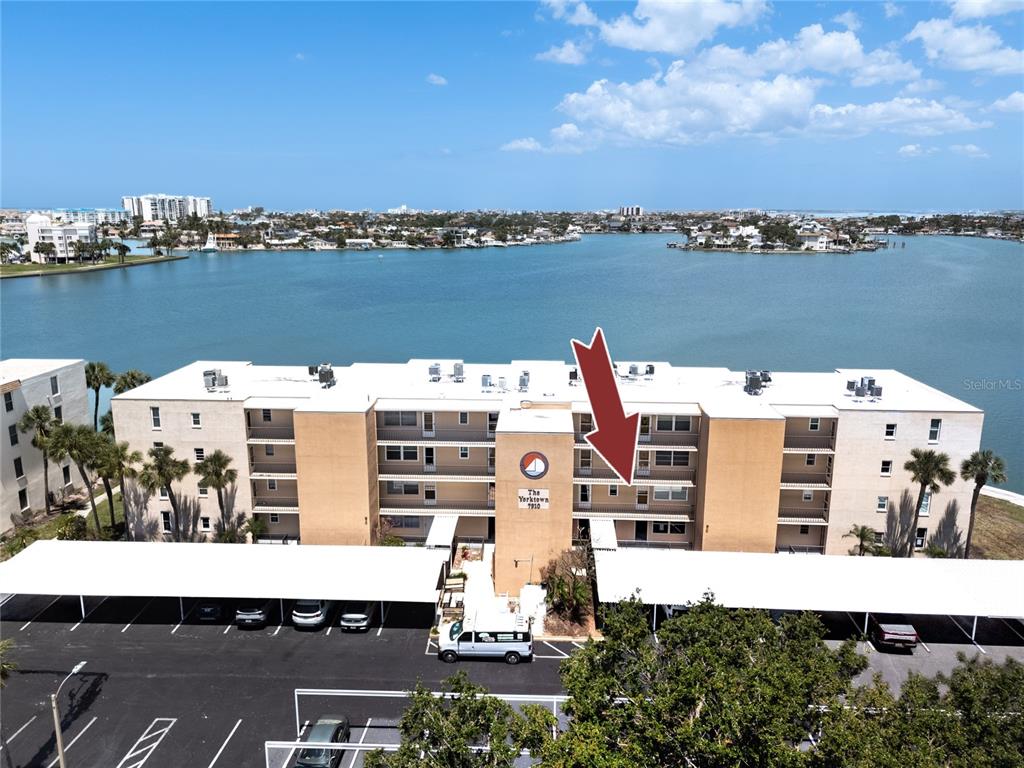 An aerial view shows a multi-story apartment building with a parking lot and waterfront views — 7910 SUN ISLAND DRIVE S UNIT 203, SOUTH PASADENA, FL