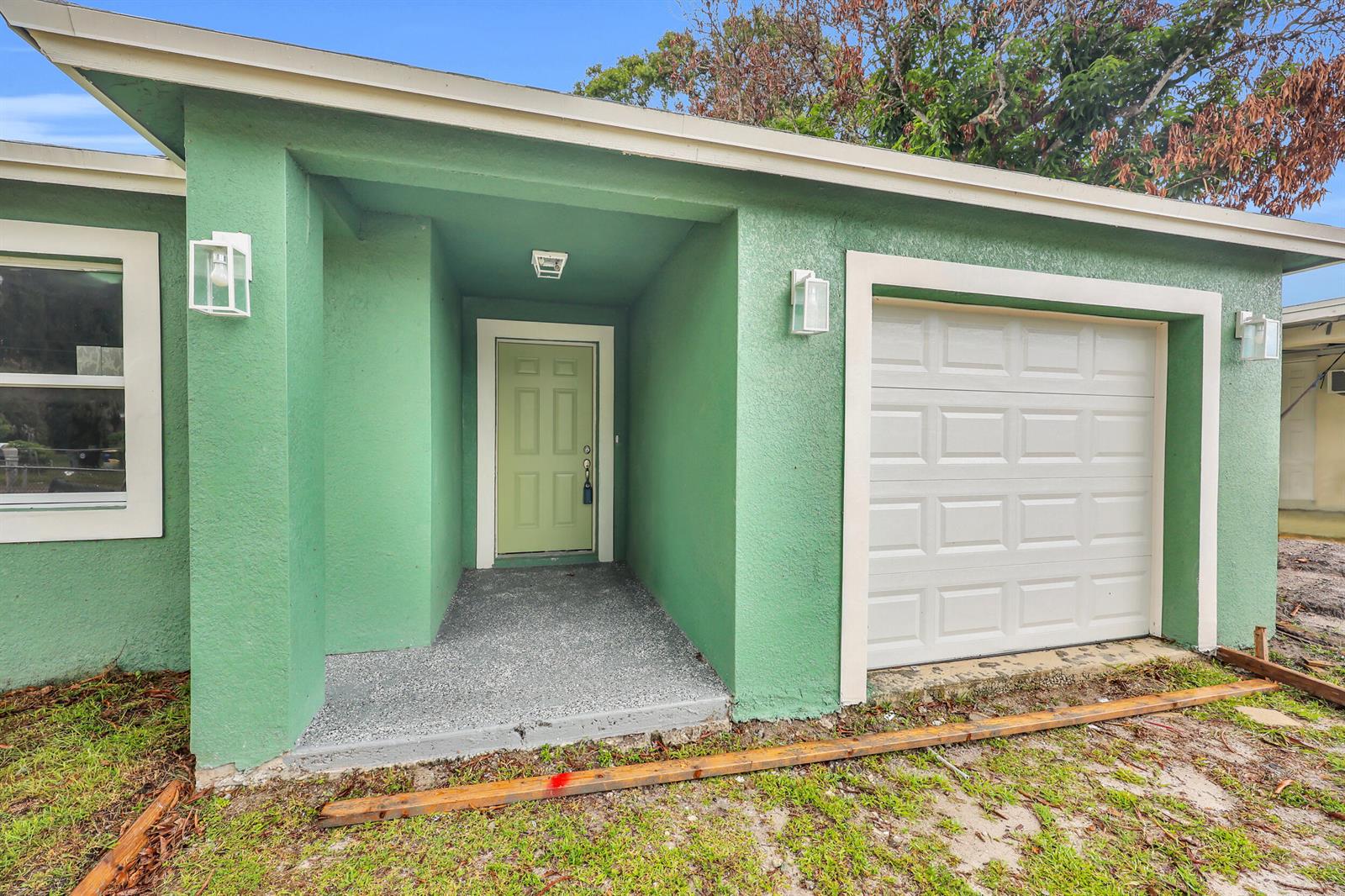 A green house exterior features a recessed front door, a garage, and outdoor lighting fixtures — 2002 Avenue M, Fort Pierce FL 34950, FORT PIERCE, FL