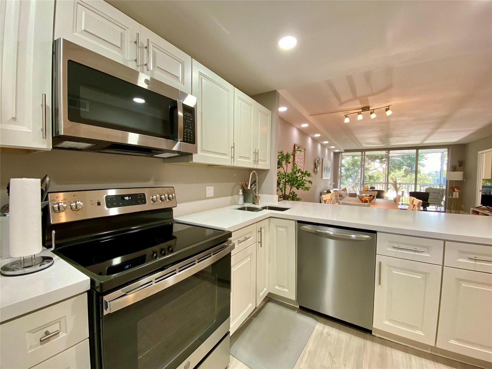 A kitchen with white cabinets, stainless steel appliances, and quartz countertops overlooks a living area — 3010 N Course Drive # 209, Pompano Beach FL 33069, POMPANO BEACH, FL