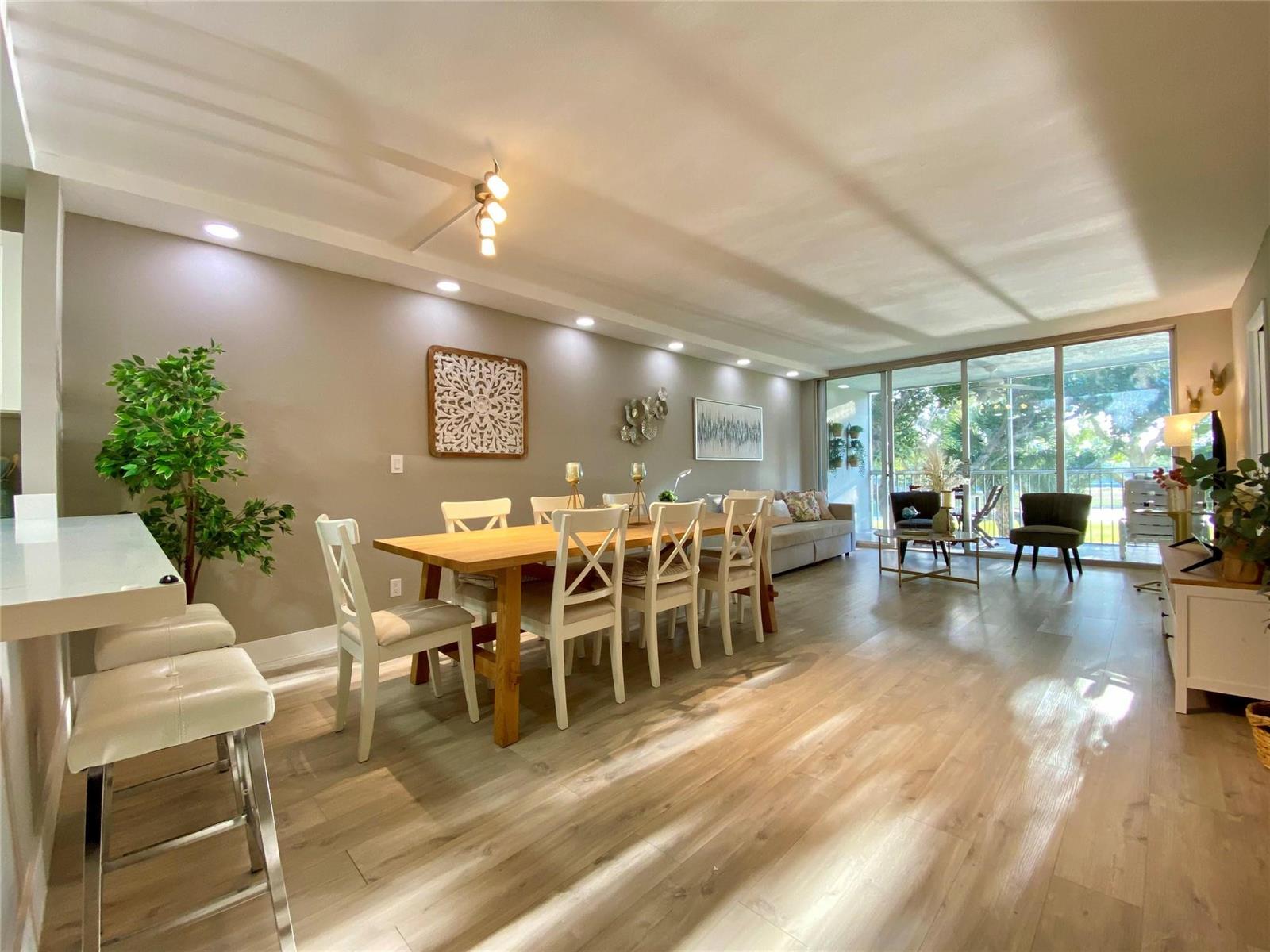 An open-concept dining and living area features light wood flooring, white furniture, and sliding glass doors — 3010 N Course Drive # 209, Pompano Beach FL 33069, POMPANO BEACH, FL