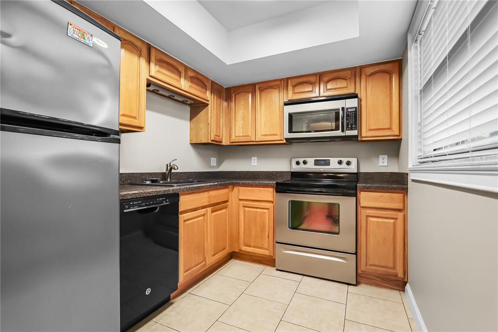 A kitchen features light wood cabinets, stainless steel appliances, dark countertops, and beige tile flooring — 3036 EASTLAND BOULEVARD UNIT E104, CLEARWATER, FL
