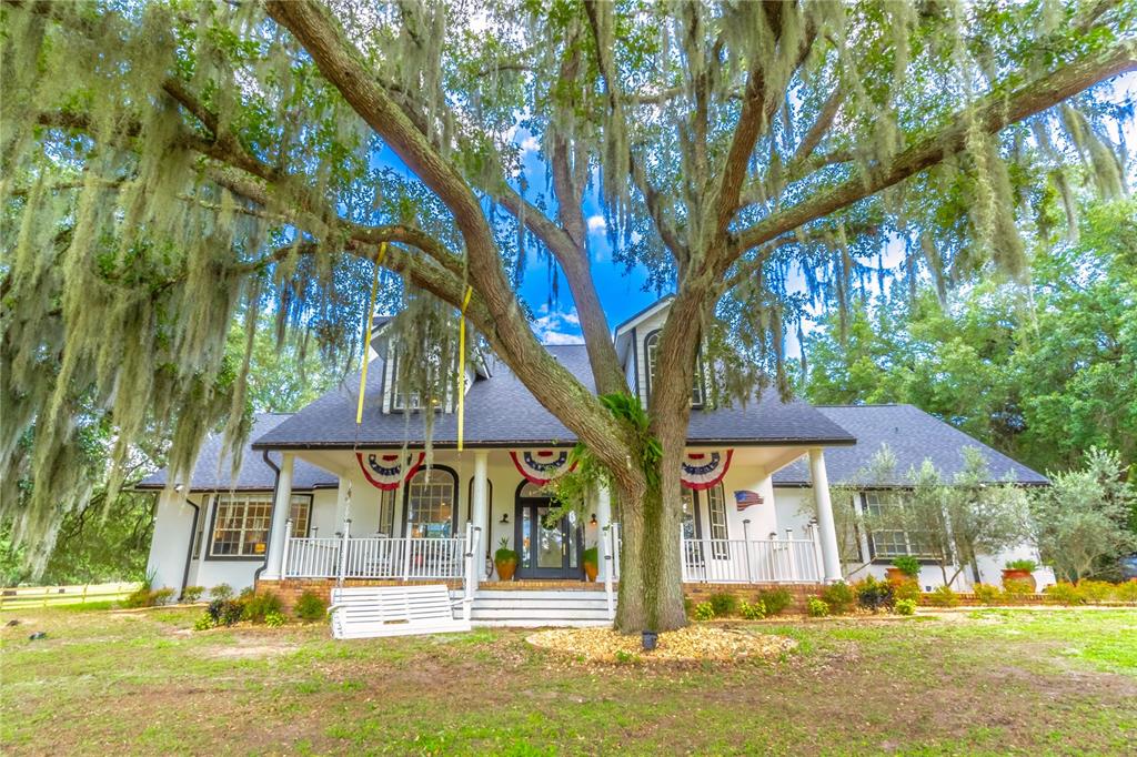 A large white house with a porch and swing sits behind a moss-covered oak tree — 23100 SE HIGHWAY 42, UMATILLA, FL