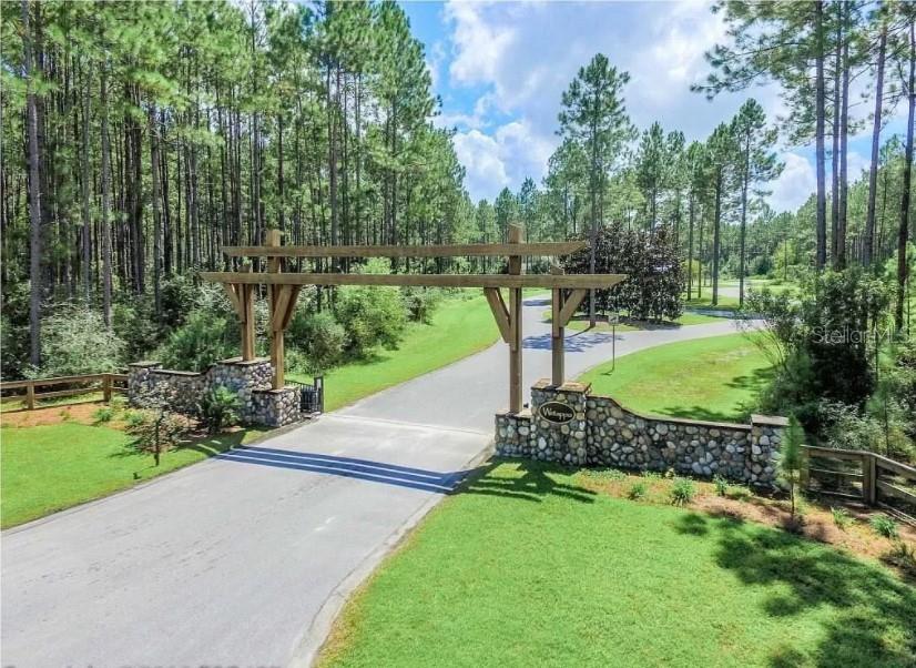 A wooden arched gate stands over a paved road entrance flanked by stone walls — CREEK PINES CIRCLE, WEWAHITCHKA, FL