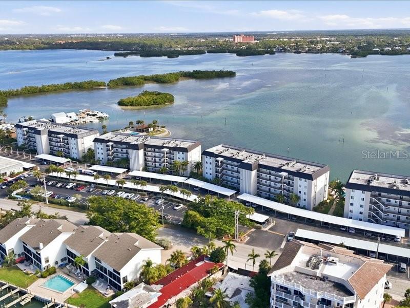 An aerial view shows several multi-story apartment buildings with covered parking lots near a bay — 8911 MIDNIGHT PASS ROAD UNIT 208, SARASOTA, FL