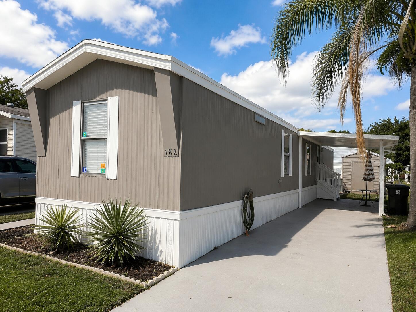 A gray mobile home with white trim, a concrete driveway, and a small palm tree — 182 Plantation Boulevard, FL 33467, FL