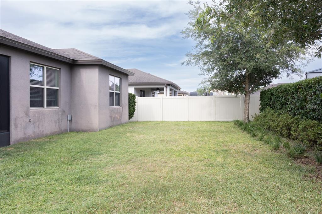 A grassy backyard features a gray stucco house exterior, a white vinyl fence, and trees — 2892 MATERA DRIVE, ST CLOUD, FL