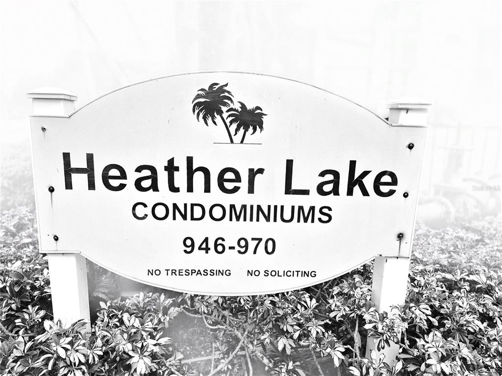 A white oval sign for Heather Lake Condominiums stands above a patch of green shrubs — 970 VIRGINIA STREET UNIT 103, DUNEDIN, FL