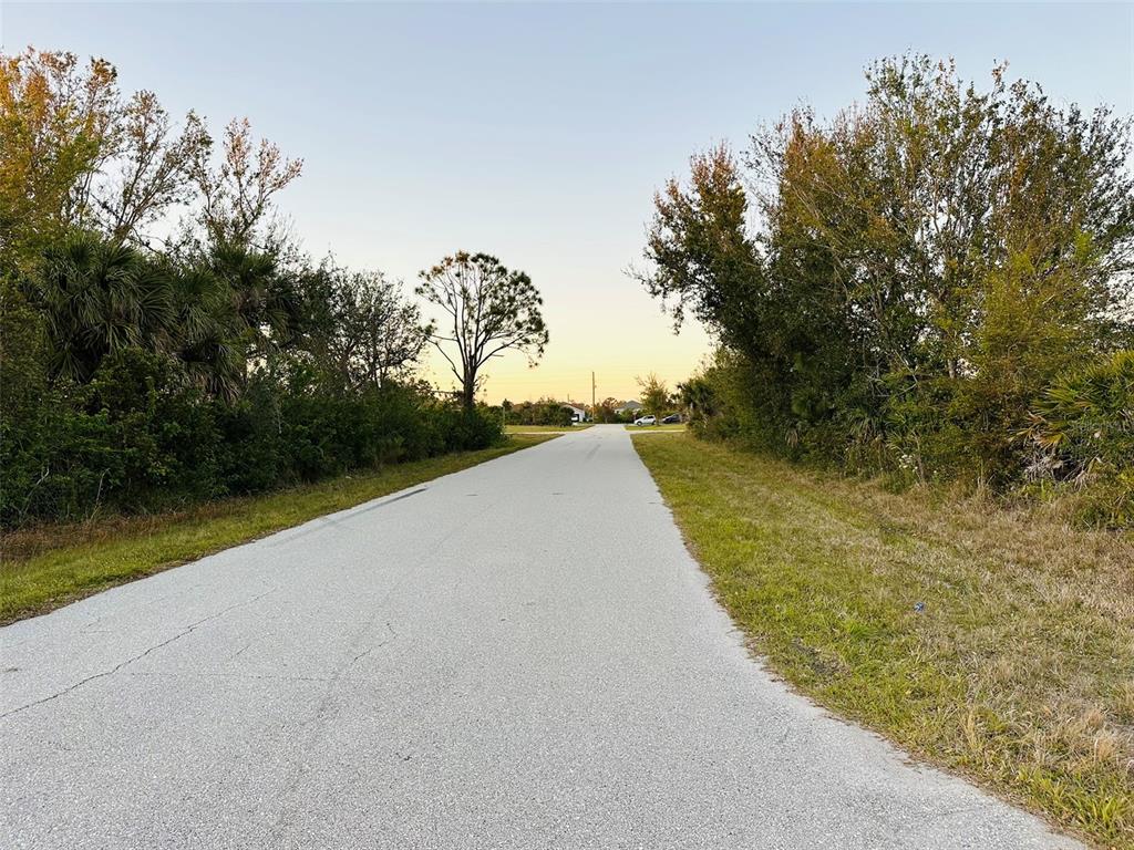 A paved road stretches between dense trees and grassy shoulders toward a distant horizon — 12204 ZITTLE AVENUE, PORT CHARLOTTE, FL