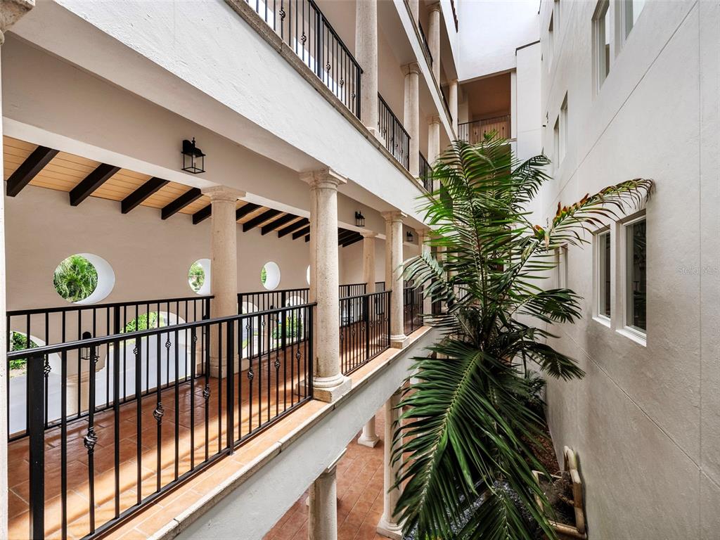 An open-air corridor with stone columns and black railings overlooks a central courtyard with palm — 11120 HACIENDA DEL MAR BOULEVARD UNIT F-205, PLACIDA, FL