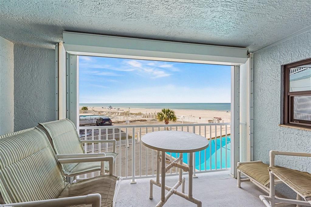 A balcony with patio furniture overlooks a swimming pool, a parking lot, and the beach — 19500 GULF BOULEVARD UNIT 102, INDIAN SHORES, FL
