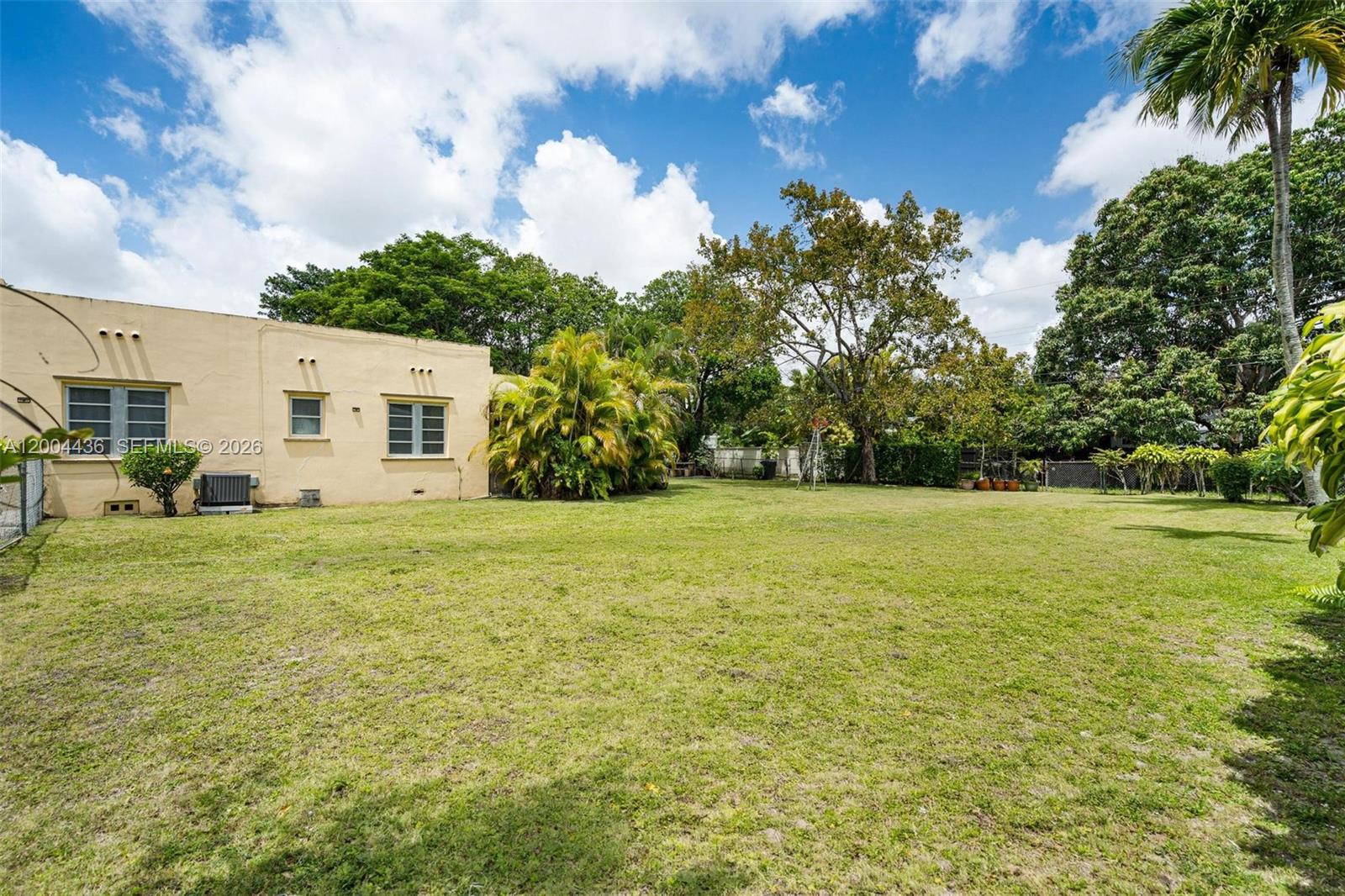 A grassy backyard features a tan stucco house, mature trees, and palm plants — 57 N Melrose Dr, Miami Springs FL 33166, MIAMI SPRINGS, FL