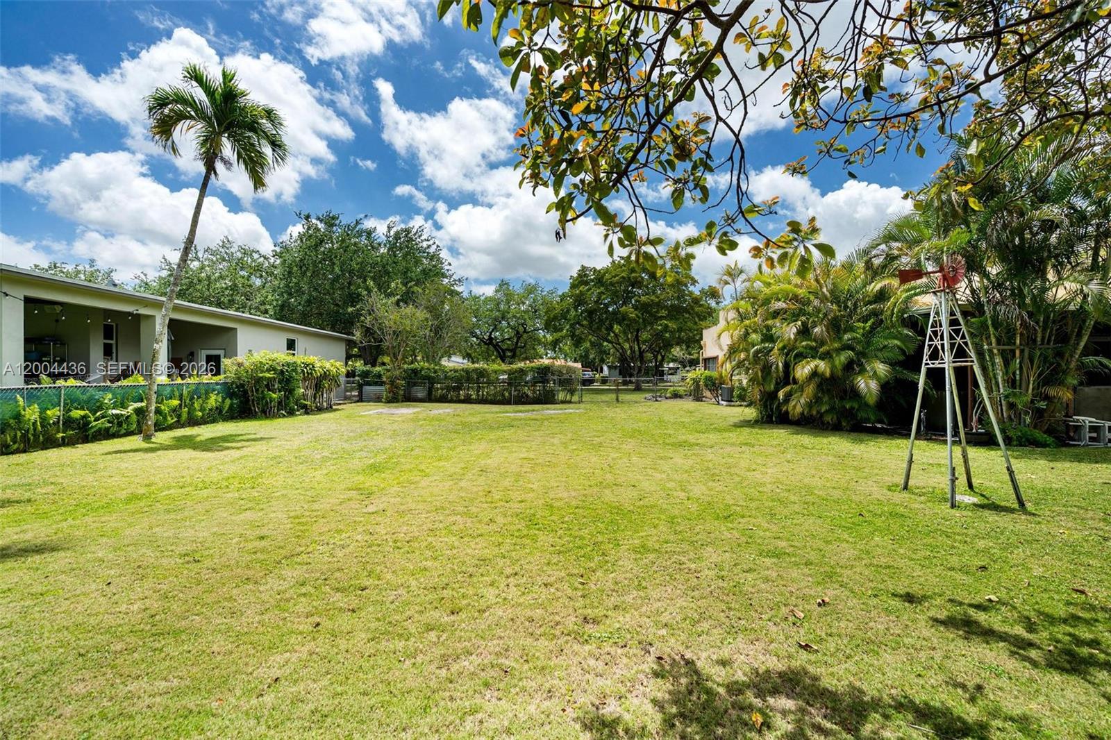 A large, grassy backyard features a palm tree, a decorative windmill, and surrounding foliage — 57 N Melrose Dr, Miami Springs FL 33166, MIAMI SPRINGS, FL