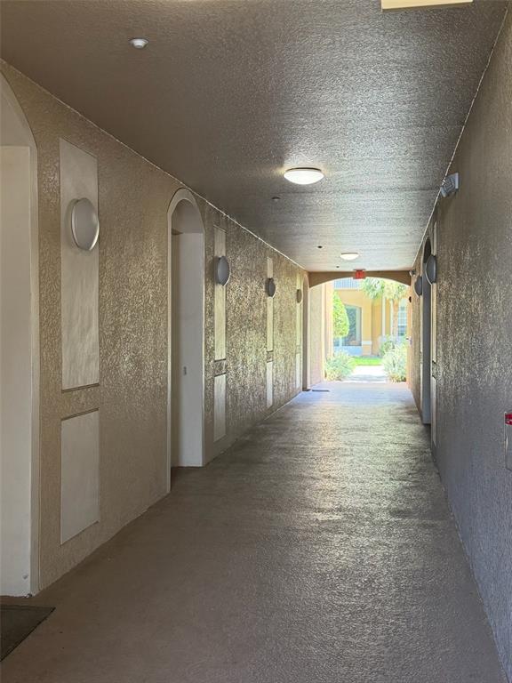 A covered outdoor hallway with textured beige walls, arched doorways, and ceiling-mounted light fixtures — 6451 OLD PARK LANE UNIT 102, ORLANDO, FL