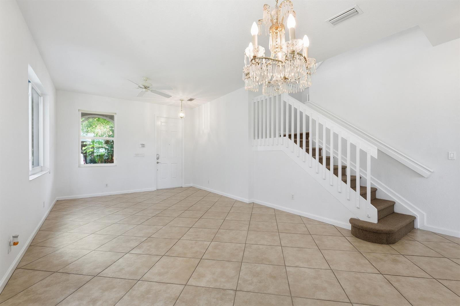 A bright, empty living room features white walls, tan tile flooring, a crystal chandelier, and stairs — 1275 Rosegate Boulevard, Riviera Beach FL 33404, RIVIERA BEACH, FL
