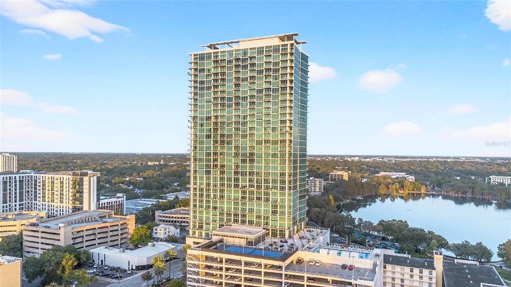 An aerial view shows a tall glass residential tower next to a lake and parking garage — 150 E ROBINSON STREET UNIT 3106, ORLANDO, FL